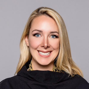 Close-up portrait of a woman with blonde hair and bright blue eyes smiling broadly, wearing a black cowl-neck garment and a gold statement necklace.